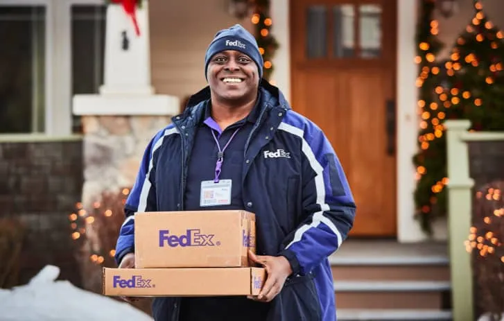 Delivery driver holding FedEx packages in front of a house during winter holiday season, smiling and wearing branded uniform.